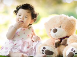 Smiling baby girl sits on grass with a plush teddy bear, enjoying a sunny day. Islamic Girls Names